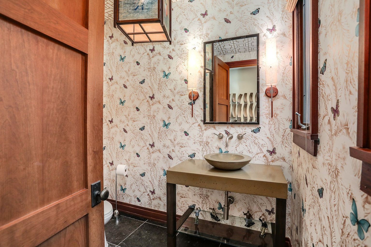 This is a charming guest bathroom featuring butterfly-themed wallpaper, a unique vessel sink on a console table, and a decorative mirror flanked by sconces. The room has a warm and inviting feel, with dark wood accents and a playful yet sophisticated design. The black tile floor adds a touch of elegance.