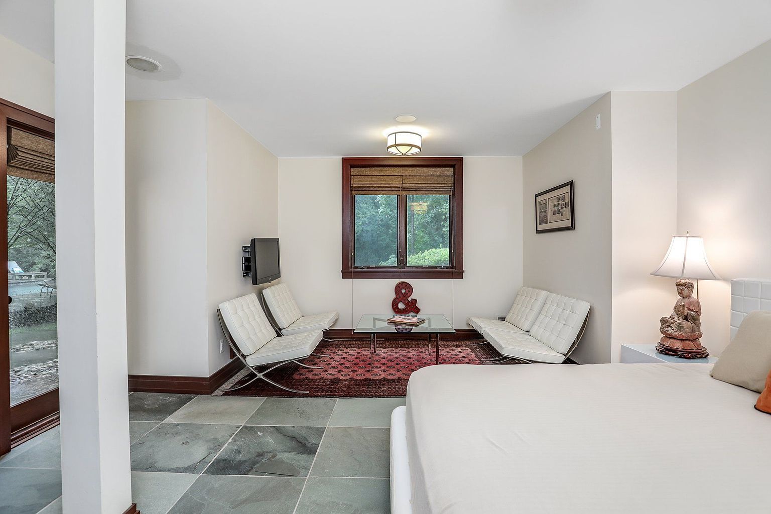 This is a well-lit primary bedroom featuring a seating area with two white Barcelona-style chairs and a glass coffee table on a red patterned rug. A flat-screen TV is mounted on the wall, and a window with wooden blinds provides natural light. The bedroom also includes a bed with white linens and a bedside table with a lamp and decorative statue, all contributing to a modern and comfortable atmosphere.