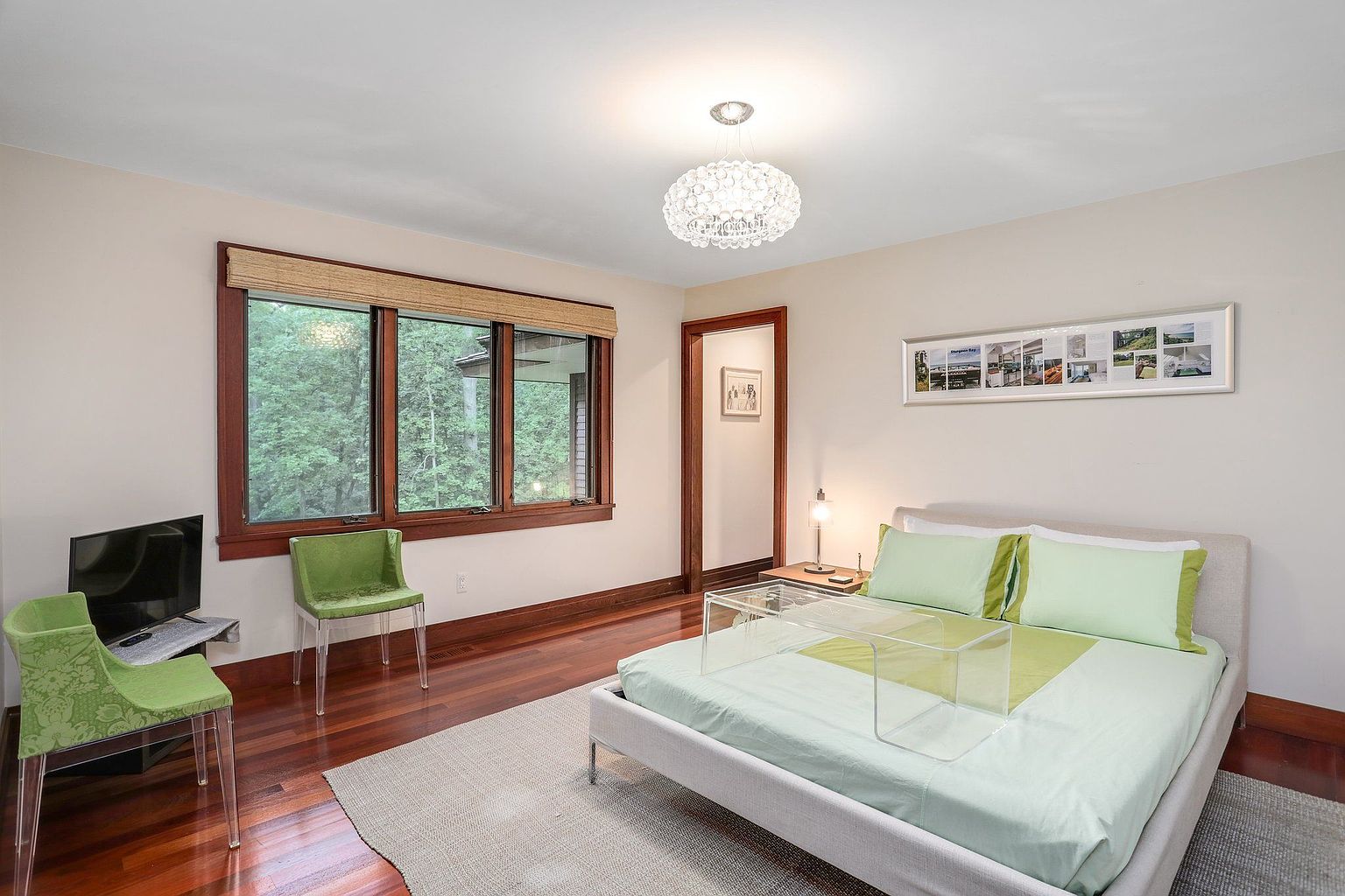 This is a well-lit primary bedroom featuring hardwood floors, a large window with a view of greenery, and a modern bed with light green bedding. The room is decorated with framed artwork and has a contemporary chandelier, creating a serene and inviting atmosphere. The presence of a TV and seating area suggests a comfortable and functional space.
