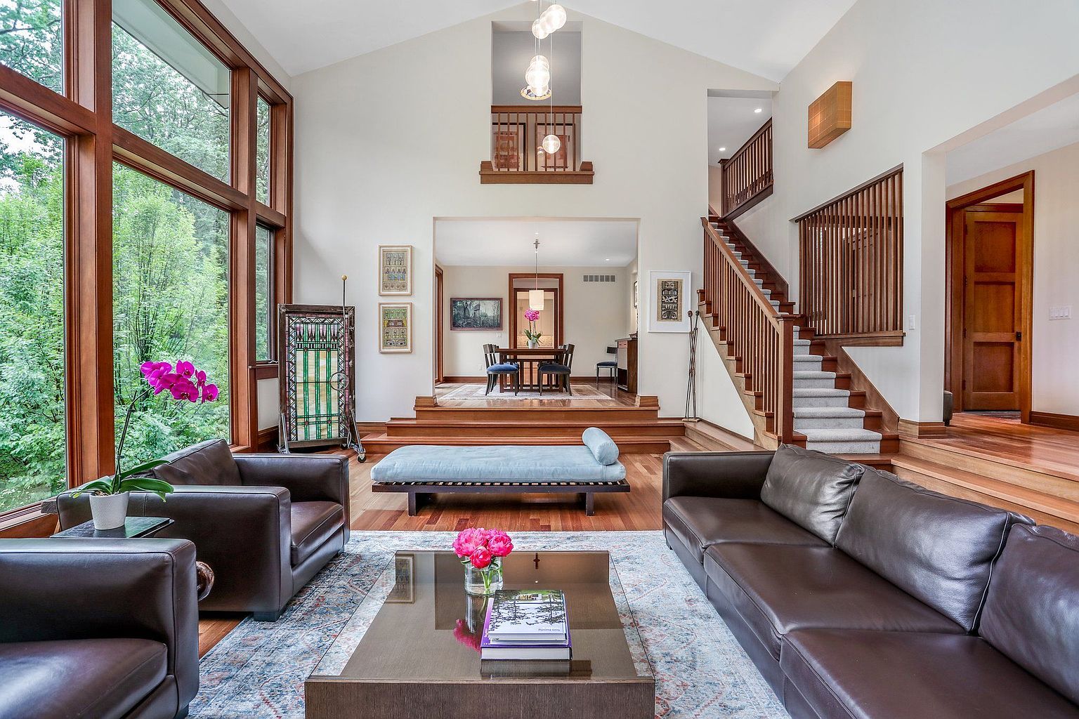 This is an interior shot of a luxurious living room featuring hardwood floors, a large window with a view of greenery, and a staircase leading to an upper level. The room is furnished with leather sofas, a coffee table, and a daybed, creating a comfortable and elegant atmosphere. The perspective is from the front of the room, capturing the depth and architectural details.
