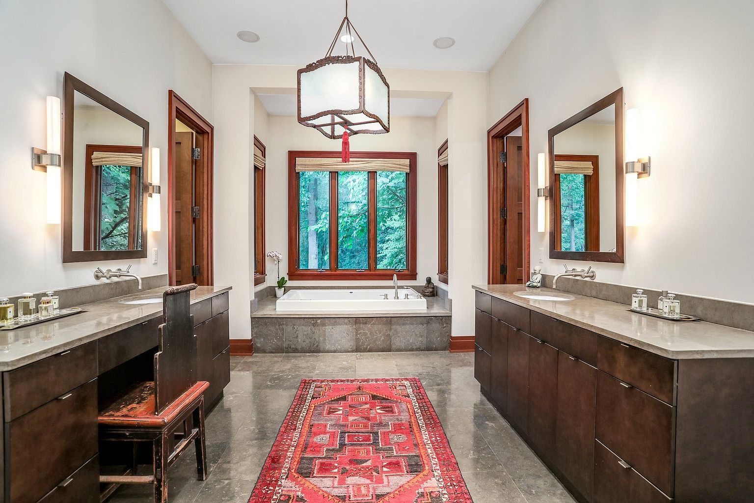 This is a well-lit primary bathroom featuring dual vanities with dark wood cabinetry and light countertops. A large window above a soaking tub provides a view of lush greenery, enhancing the spa-like atmosphere. A red oriental rug adds a pop of color to the gray tile floor, and unique lighting fixtures complete the elegant design.