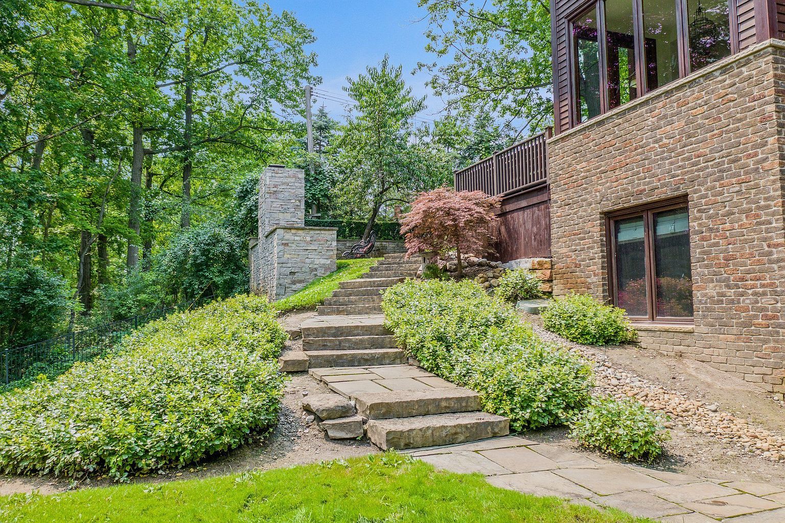 This image showcases a beautifully landscaped yard with stone steps leading up a gentle slope. Lush greenery flanks the steps, and a stone structure is visible at the top, possibly a fireplace or architectural feature. The brick exterior of the house is also visible, adding to the overall charm and appeal of the property.