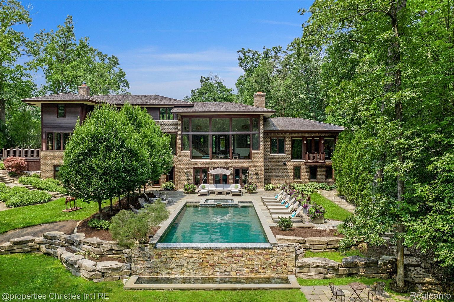This is a rear exterior view of a luxurious home featuring a stunning pool and spa area. The pool is surrounded by a stone patio with lounge chairs and an umbrella-covered seating area. The house itself is a blend of brick and wood, with large windows offering views of the interior, all set against a backdrop of lush greenery and mature trees.