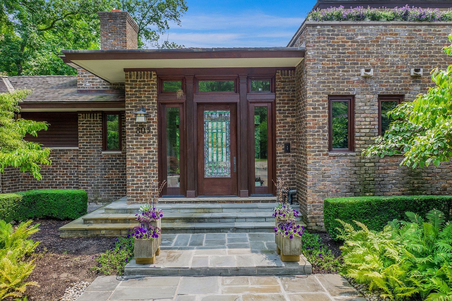 This image showcases the entryway of a brick home, featuring a prominent wooden door with decorative glass panels, flanked by sidelights and topped with transom windows. Stone steps lead up to the entrance, which is adorned with potted purple flowers on either side. The surrounding landscaping includes manicured hedges and ferns, creating a welcoming and sophisticated curb appeal.