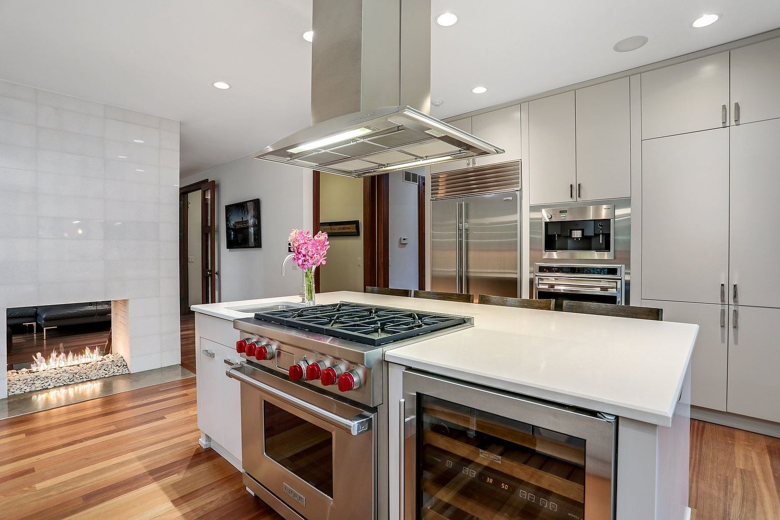 This is a well-lit, modern kitchen featuring stainless steel appliances, including a large refrigerator, oven, and wine cooler. The kitchen island has a white countertop and a built-in stovetop. The hardwood floors add warmth to the space, and a fireplace is visible in the background, creating a cozy atmosphere.