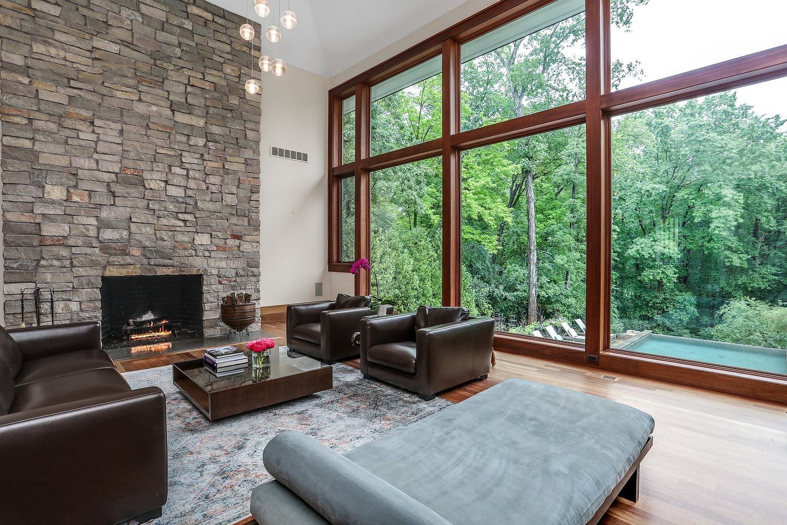 This is an interior shot of a luxurious living room featuring a stone fireplace, large windows with a view of lush greenery and a pool, and modern furniture. The room is well-lit and spacious, creating a comfortable and elegant atmosphere. The perspective is from a medium distance, capturing the entire living space and its connection to the outdoor environment.