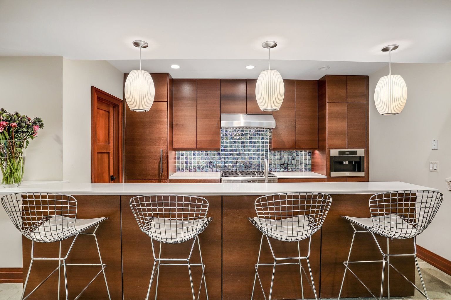 This is a well-lit kitchen featuring modern, dark wood cabinetry and a white countertop bar area with four metal bar stools. The backsplash is a mosaic tile pattern, and pendant lights hang above the bar. The kitchen also includes stainless steel appliances and a built-in coffee maker, creating a sleek and functional space.
