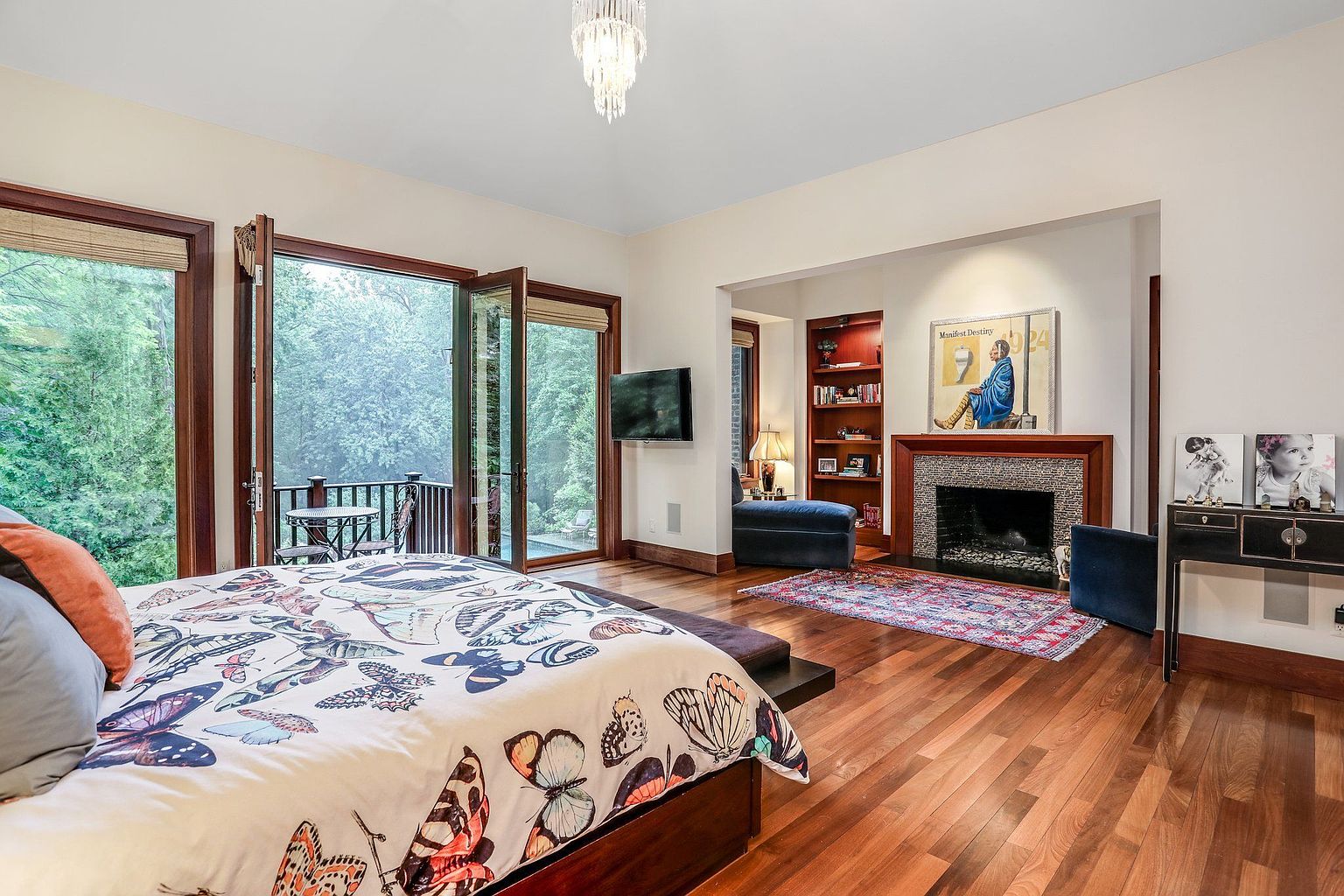 This is a primary bedroom featuring a bed with a butterfly-themed comforter, a fireplace, and access to a balcony. The room has hardwood floors, a dark wood trim, and a chandelier. The style is luxurious and comfortable, with a focus on natural light and outdoor views.