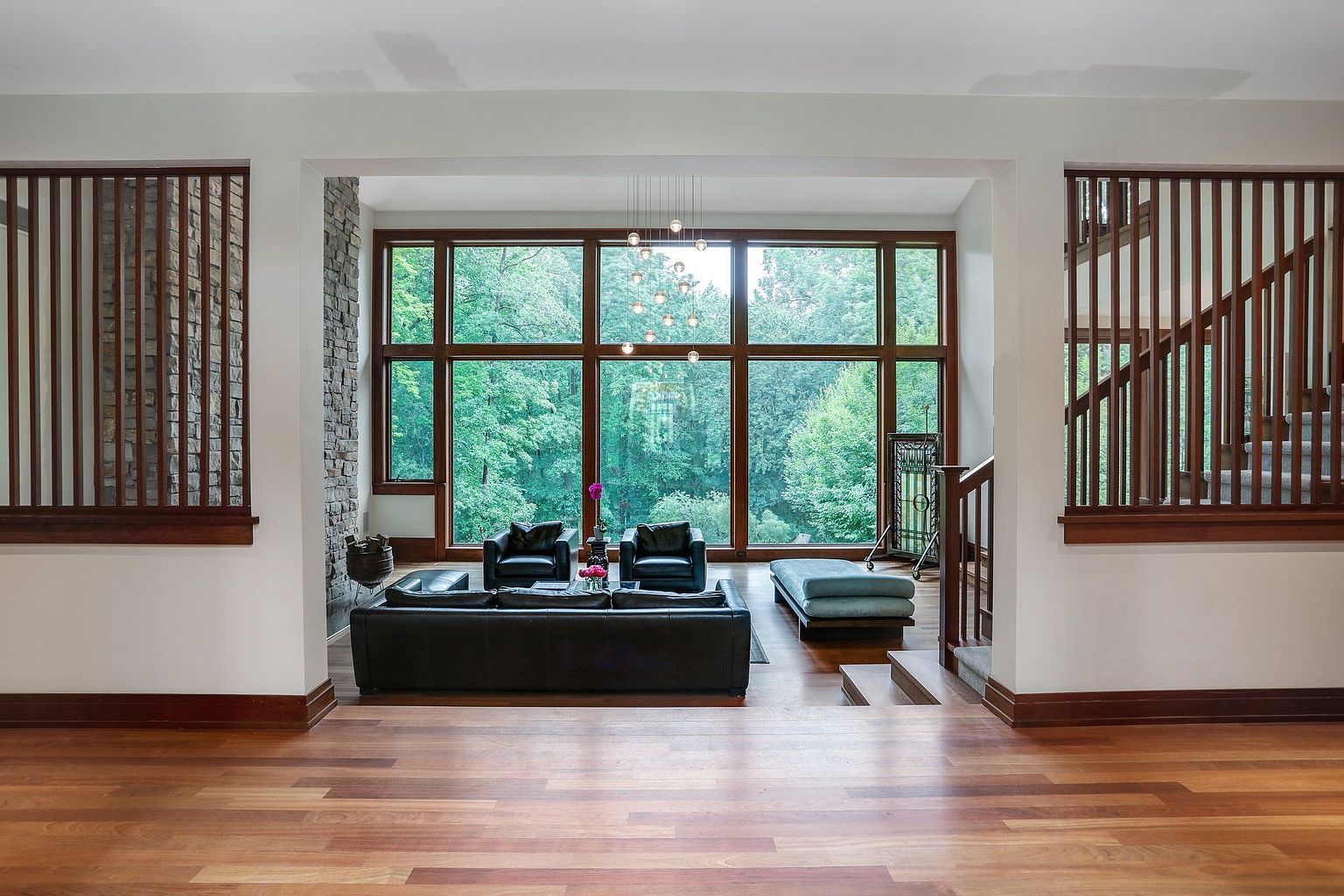 This is an interior shot of a luxurious living room featuring a large window with a view of lush greenery. The room is furnished with a black leather sofa, armchairs, and a light blue ottoman. The space is well-lit and features hardwood floors, creating a warm and inviting atmosphere.