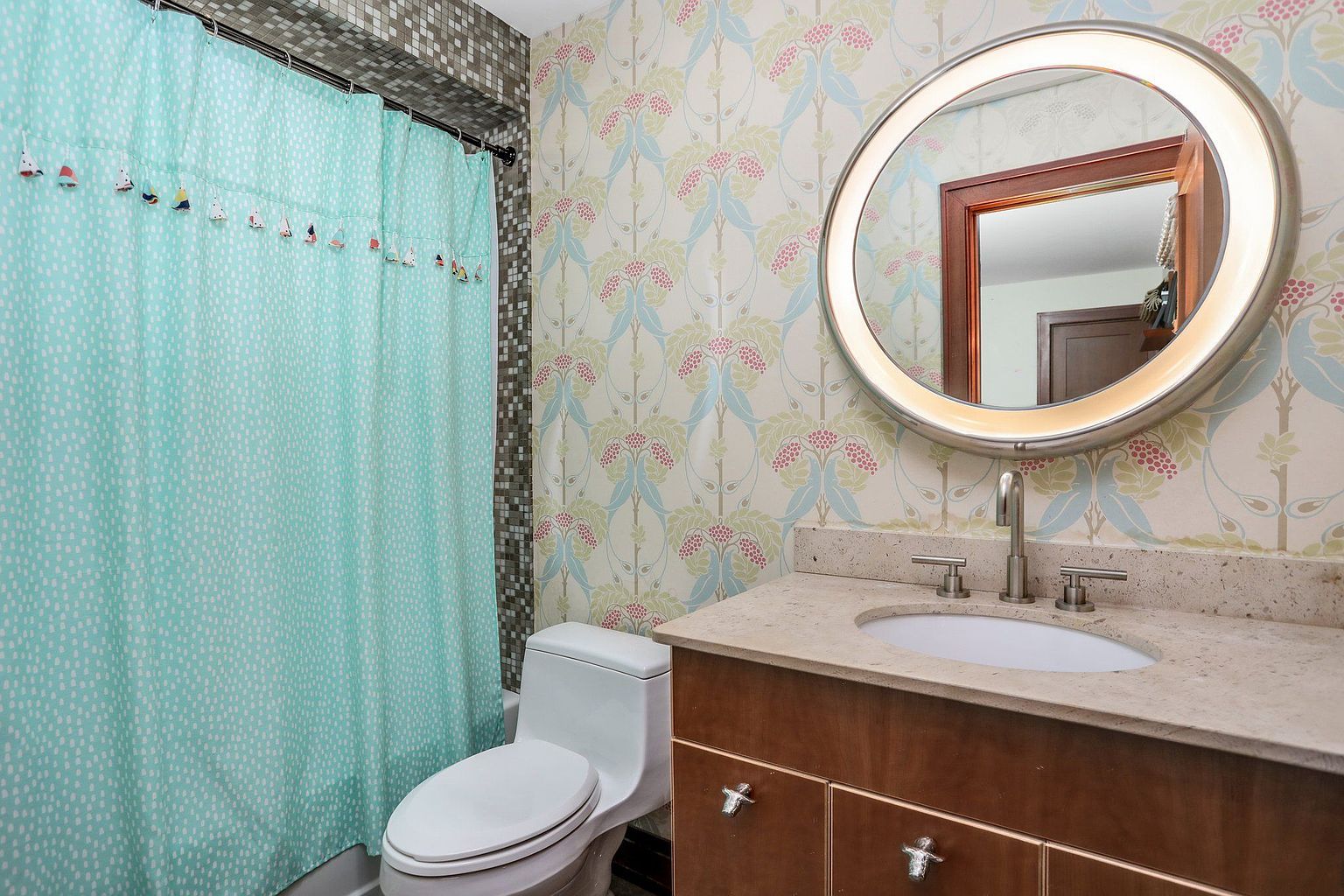 This is a bathroom featuring a toilet, a vanity with a sink, and a shower with a patterned curtain. The wallpaper has a floral design, and the mirror is round with integrated lighting. The overall impression is a clean and well-maintained space.