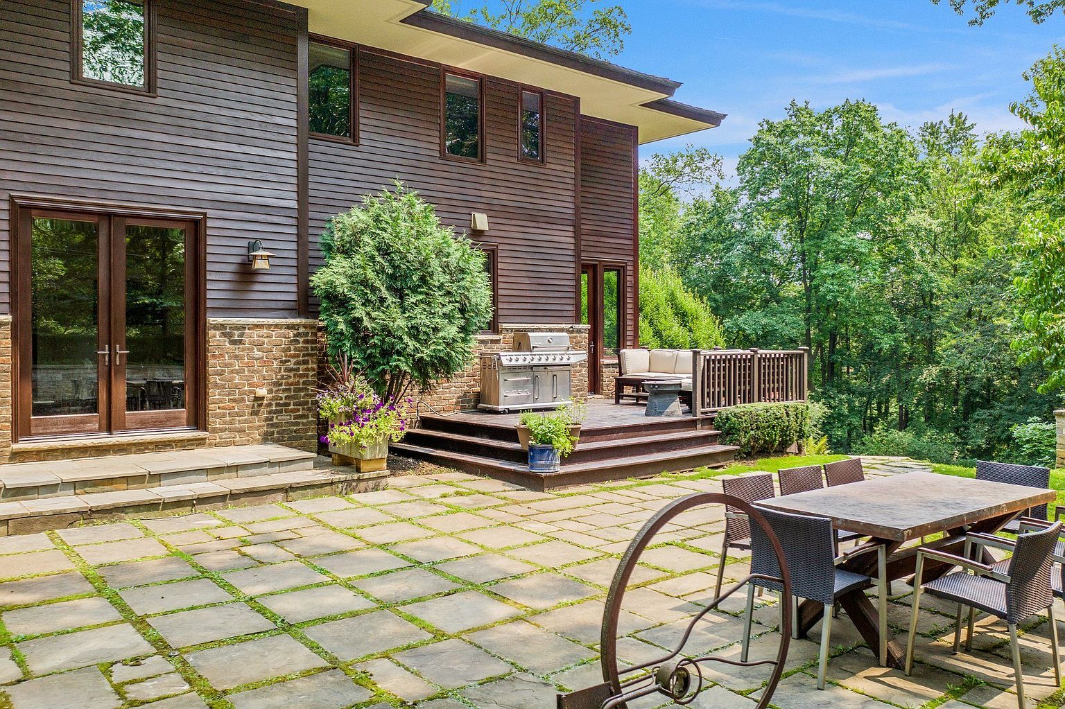 This image showcases a beautifully designed outdoor living space, featuring a stone patio with lush greenery between the pavers, a wooden deck with a built-in grill and comfortable seating, and a dining table with chairs. The exterior of the house is clad in dark wood siding with brick accents, creating a warm and inviting atmosphere. The surrounding trees provide privacy and a sense of tranquility.