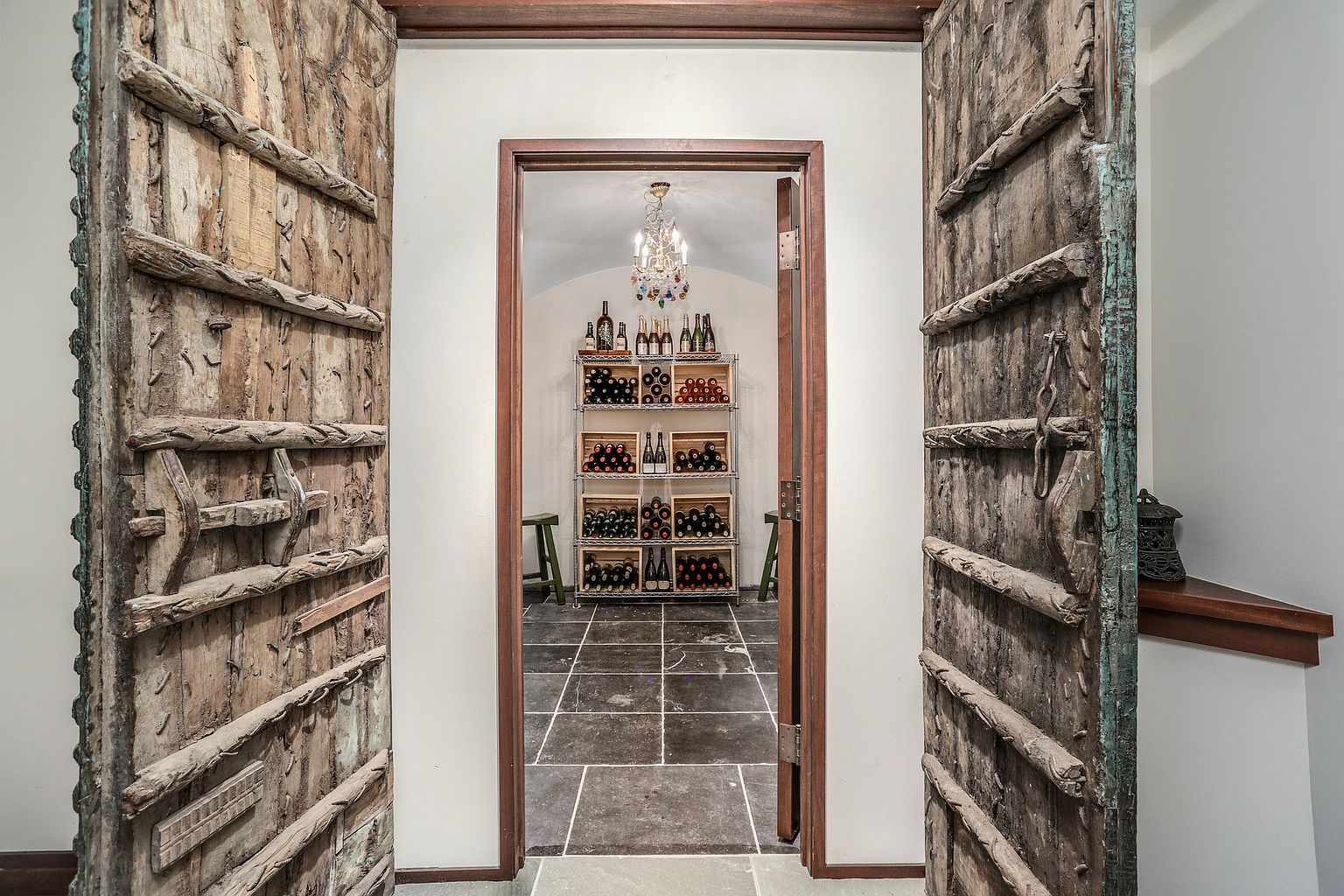 This image showcases a wine cellar viewed through a doorway framed by rustic wooden doors. Inside, a metal rack displays numerous wine bottles, illuminated by a chandelier. The floor is tiled, and the overall impression is one of old-world charm and sophistication.