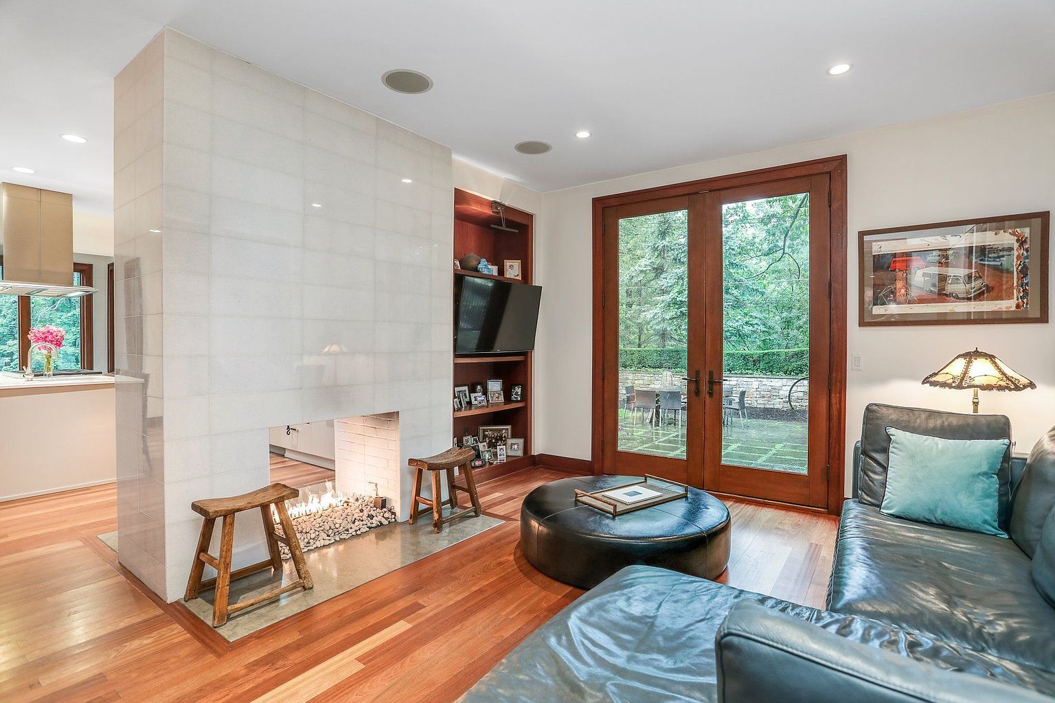 This is an interior shot of a living room featuring a modern fireplace with a white tiled facade and a hearth filled with decorative stones. The room has hardwood floors, a dark leather sofa with a light blue pillow, and a round leather ottoman. Double doors lead to an outdoor patio, and built-in shelves with a TV are visible to the right of the fireplace.