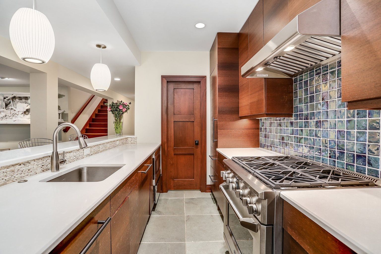 This is a view of a modern kitchen featuring wood cabinetry, stainless steel appliances, and white countertops. The backsplash is a mosaic tile with blue and green hues. The kitchen also features a stainless steel sink and faucet, as well as pendant lighting.