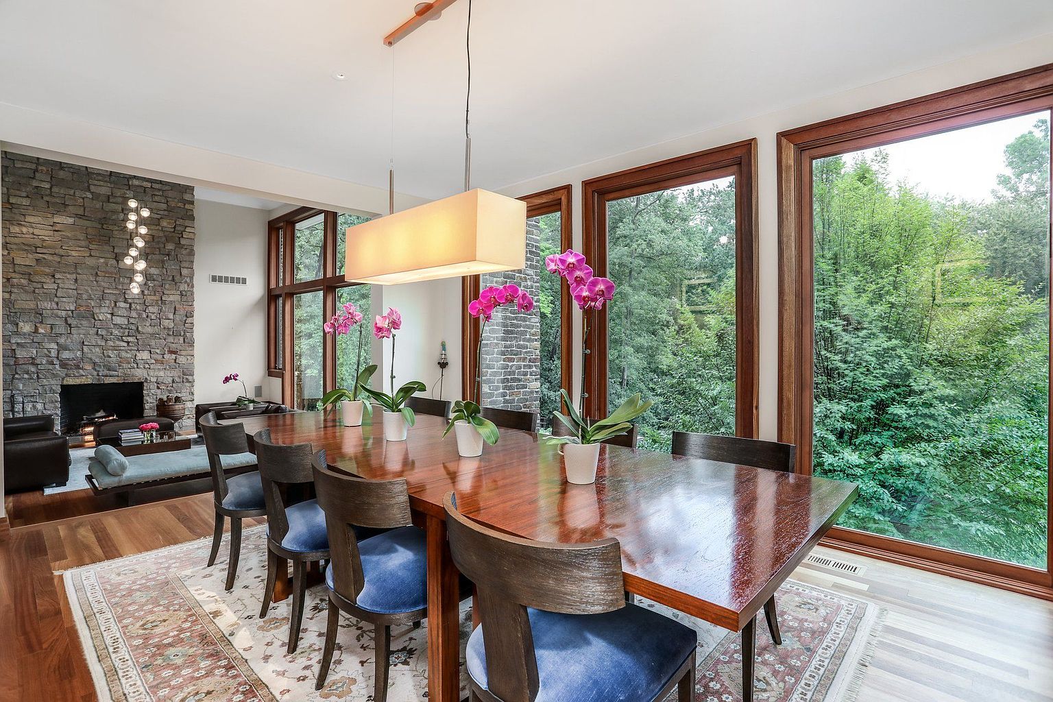This is an interior shot of a dining room featuring a long wooden table with blue upholstered chairs. The room is well-lit by a rectangular pendant light fixture and natural light streaming through large windows that offer a view of lush greenery. A stone fireplace is visible in the background, adding a touch of rustic charm to the modern space.