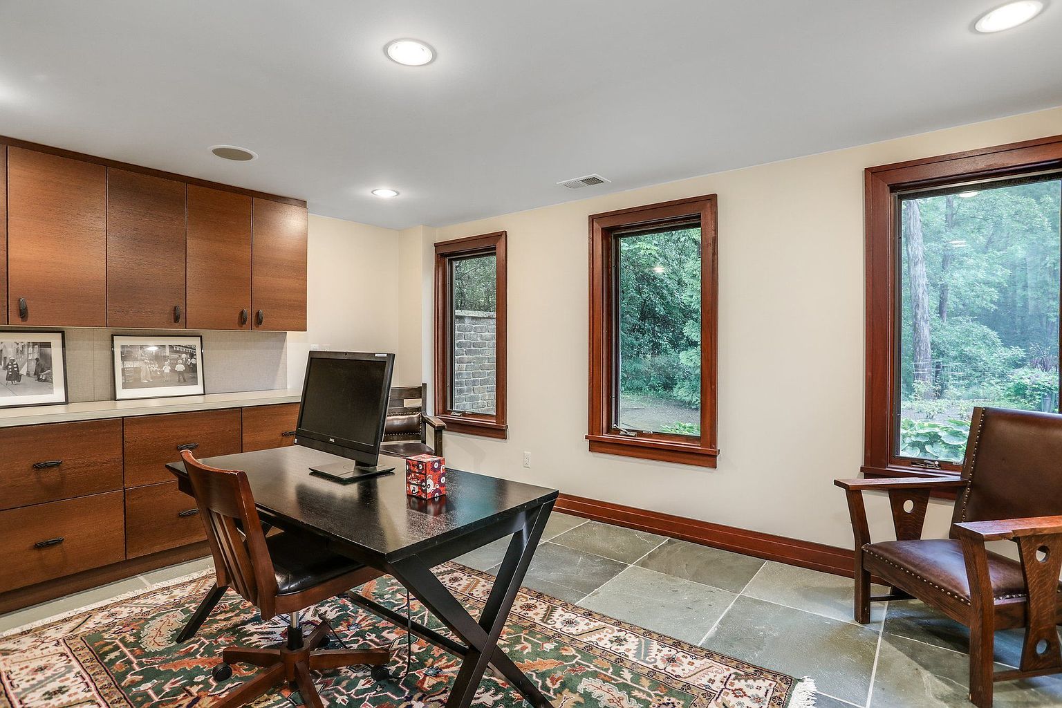 This is an interior shot of a home office featuring built-in wooden cabinets and drawers along one wall, providing ample storage. A dark-colored desk with a computer monitor sits in the center of the room, complemented by a wooden chair. Three windows offer natural light and views of the outdoors, while a patterned rug adds warmth and character to the space.