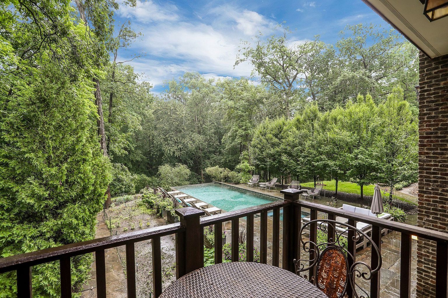 This image showcases a serene outdoor living space with a view of a luxurious pool and lush greenery. The patio features a wicker table and chair set, complemented by a dark wood railing. The pool area includes lounge chairs and umbrellas, creating an inviting atmosphere for relaxation and entertainment.