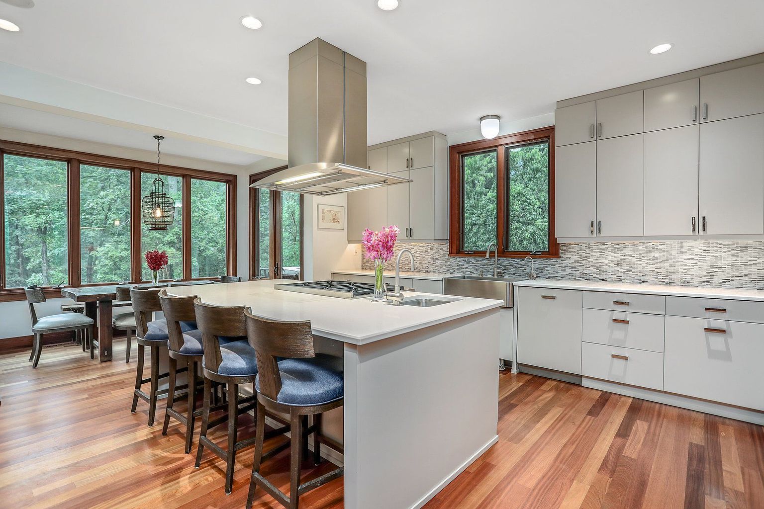 This is a bright and modern kitchen featuring light gray cabinetry, stainless steel appliances, and a large island with a white countertop. The kitchen has a large window overlooking a wooded area, providing natural light and a scenic view. The hardwood floors add warmth to the space, and the overall design is clean and contemporary, perfect for cooking and entertaining.
