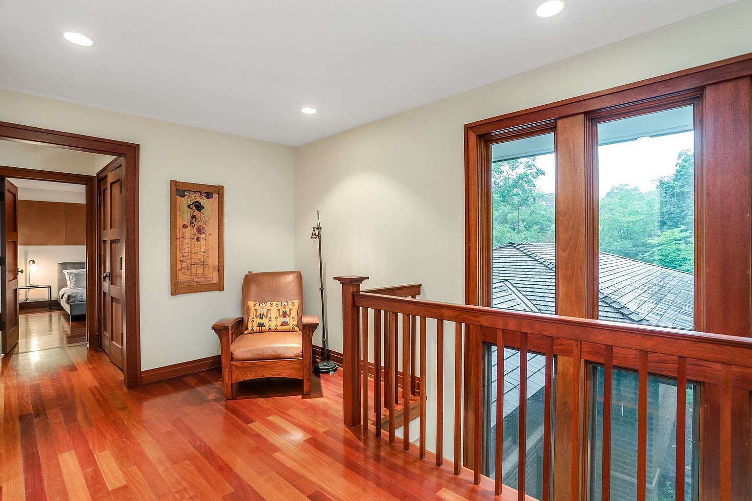 This interior shot showcases a hallway with rich hardwood flooring and wood trim. A cozy reading nook is created with a leather armchair and a framed artwork on the wall. Natural light floods the space through a large window, offering a view of the outdoors.