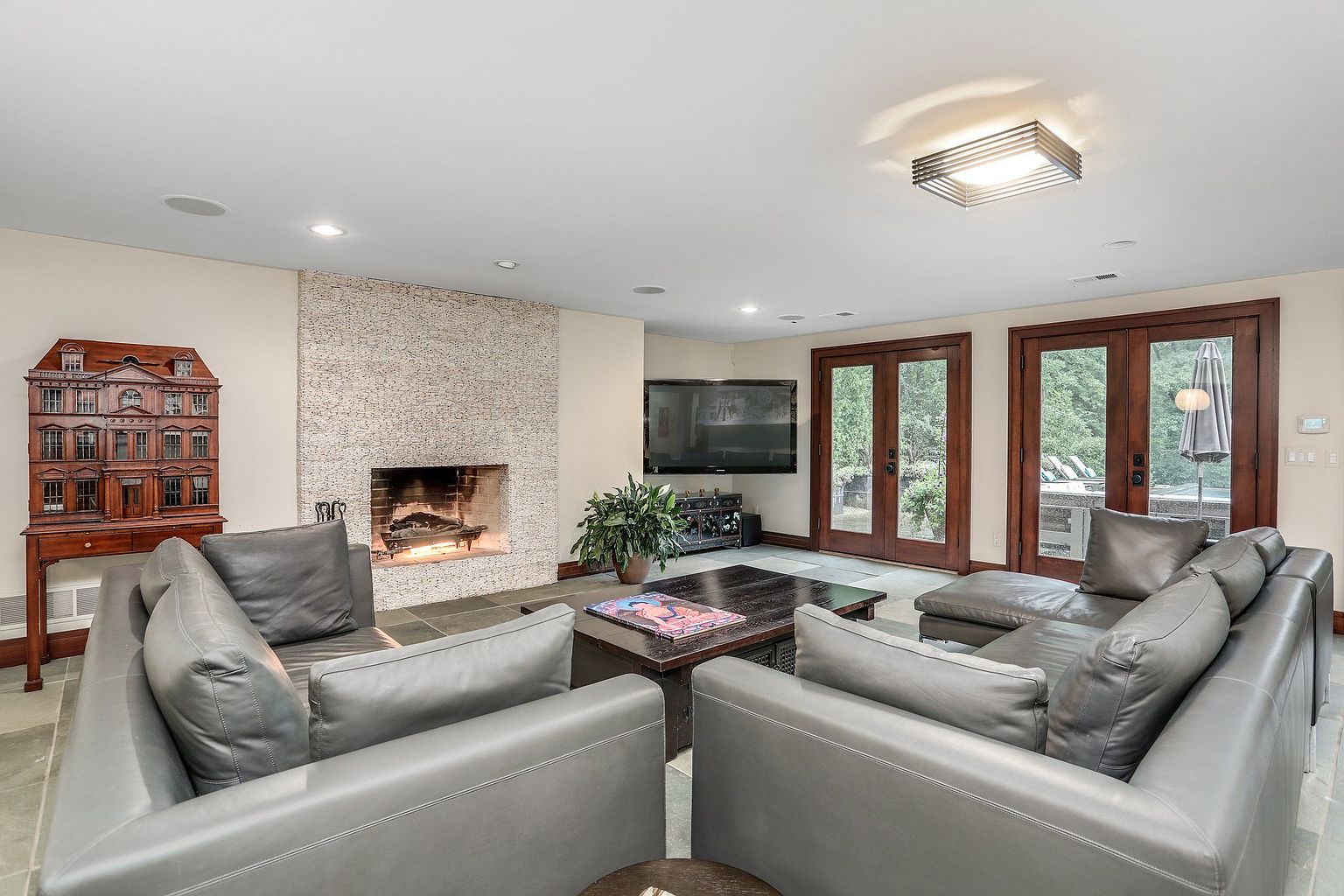 This is an interior shot of a living room featuring two gray leather sofas facing each other, a dark wood coffee table, and a fireplace with a stone surround. A large television is mounted on the wall, and double doors lead to an outdoor area. The room is well-lit with recessed lighting and a modern light fixture, creating a comfortable and inviting atmosphere.
