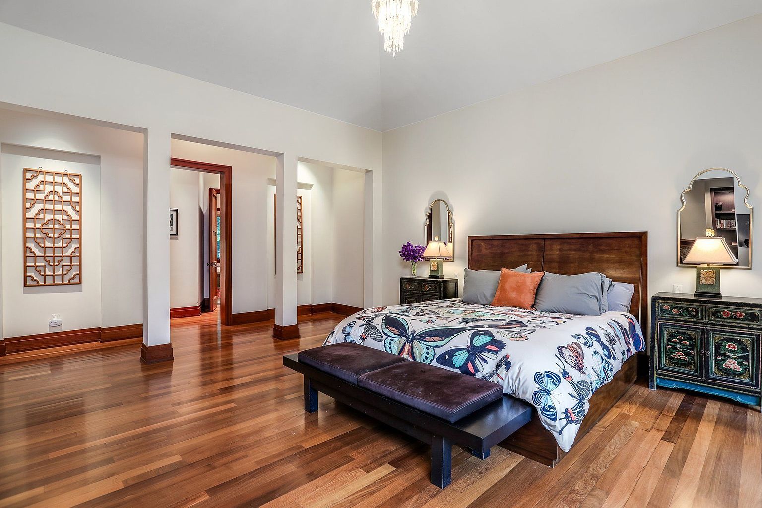 This is a primary bedroom featuring hardwood floors, a large bed with a butterfly-themed comforter, and dark wood furniture. The room has an open layout with columns defining the space, and a chandelier hangs from the ceiling. The style is a blend of traditional and eclectic, creating a comfortable and inviting atmosphere.