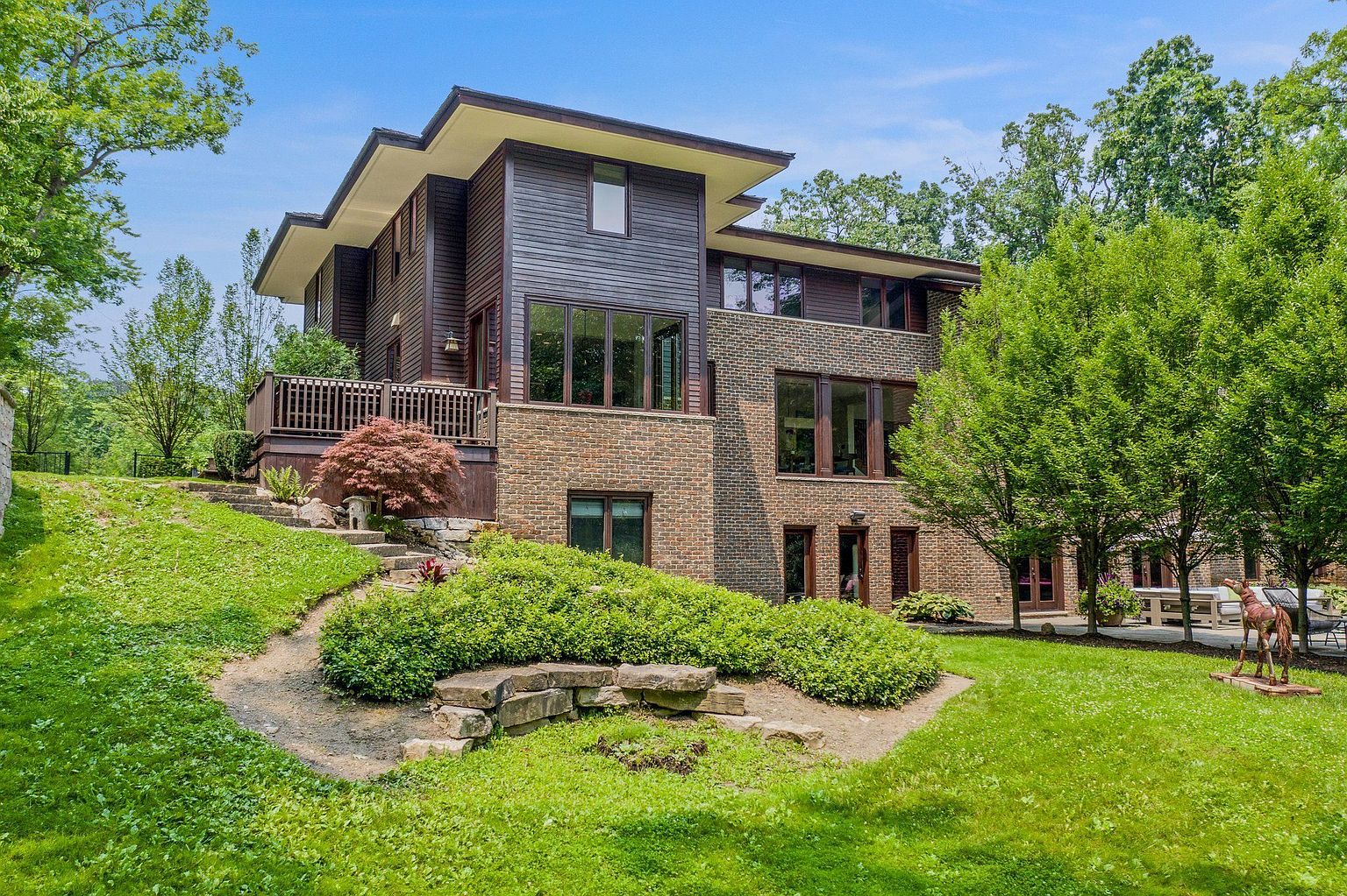 This is a front exterior view of a modern, multi-level home featuring a combination of wood siding and brick facade. The house is nestled on a well-maintained lawn with landscaped garden beds and mature trees. A wooden deck is visible on the side, adding to the property's appeal.