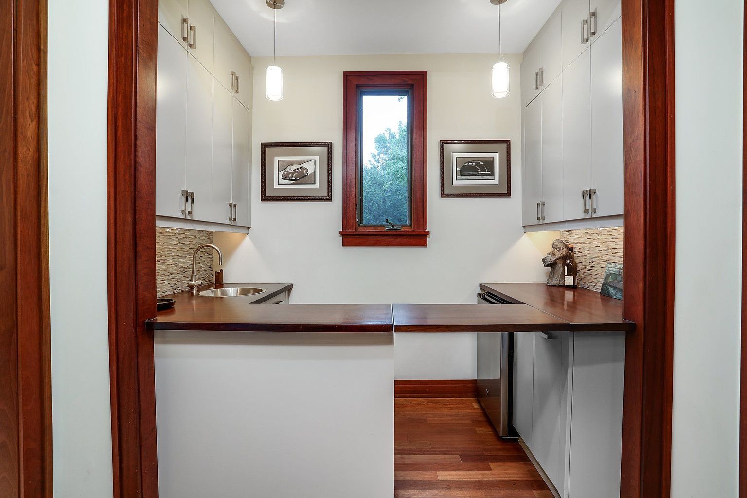This interior shot showcases a compact kitchen area with light gray cabinetry and a dark wood countertop that extends into a bar-like surface. A small window provides natural light, complemented by pendant lights, and framed artwork adds a touch of personality. The space is framed by dark wood trim, creating a warm and inviting atmosphere.