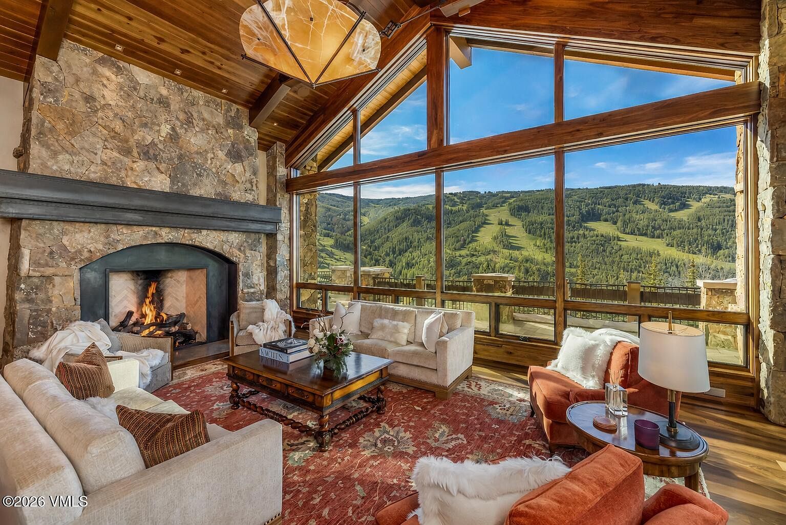This expansive living room features a dramatic vaulted wood-paneled ceiling and a prominent stone-clad chimney breast housing a roaring fireplace. A wall of large windows provides an unobstructed, panoramic view of a lush, mountainous ski landscape. The space is furnished with a mix of plush neutral sofas and warm-toned armchairs arranged around a central wooden coffee table on a large, patterned area rug.