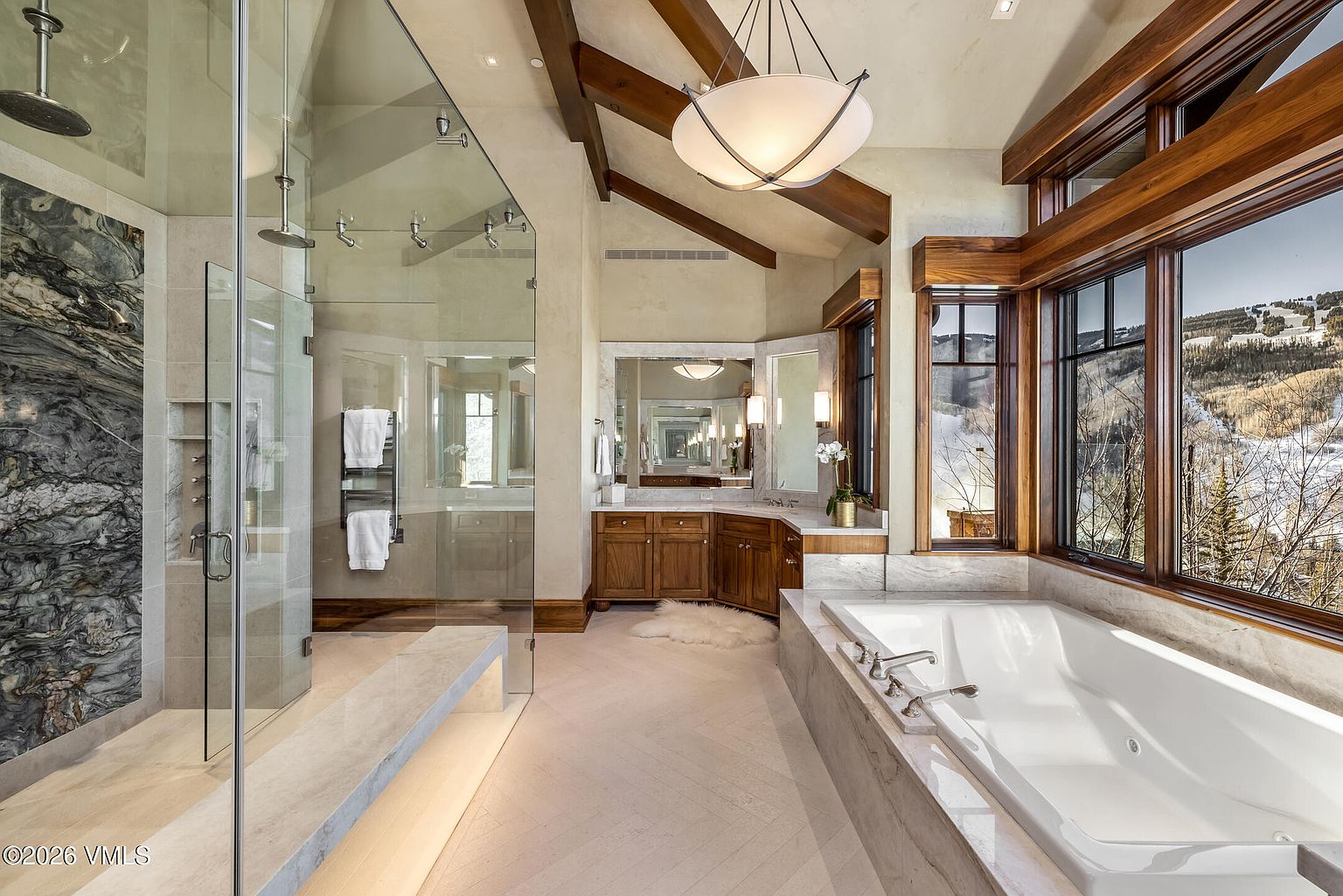 This luxurious primary bathroom features a spacious glass-enclosed walk-in shower with a striking dark stone accent wall and a separate deep soaking tub positioned beside large windows that offer scenic mountain views. The room is accented by warm wood cabinetry, exposed timber ceiling beams, and a light-toned herringbone tile floor. The perspective captures the entire layout, highlighting the blend of rustic architectural elements and modern, high-end finishes.