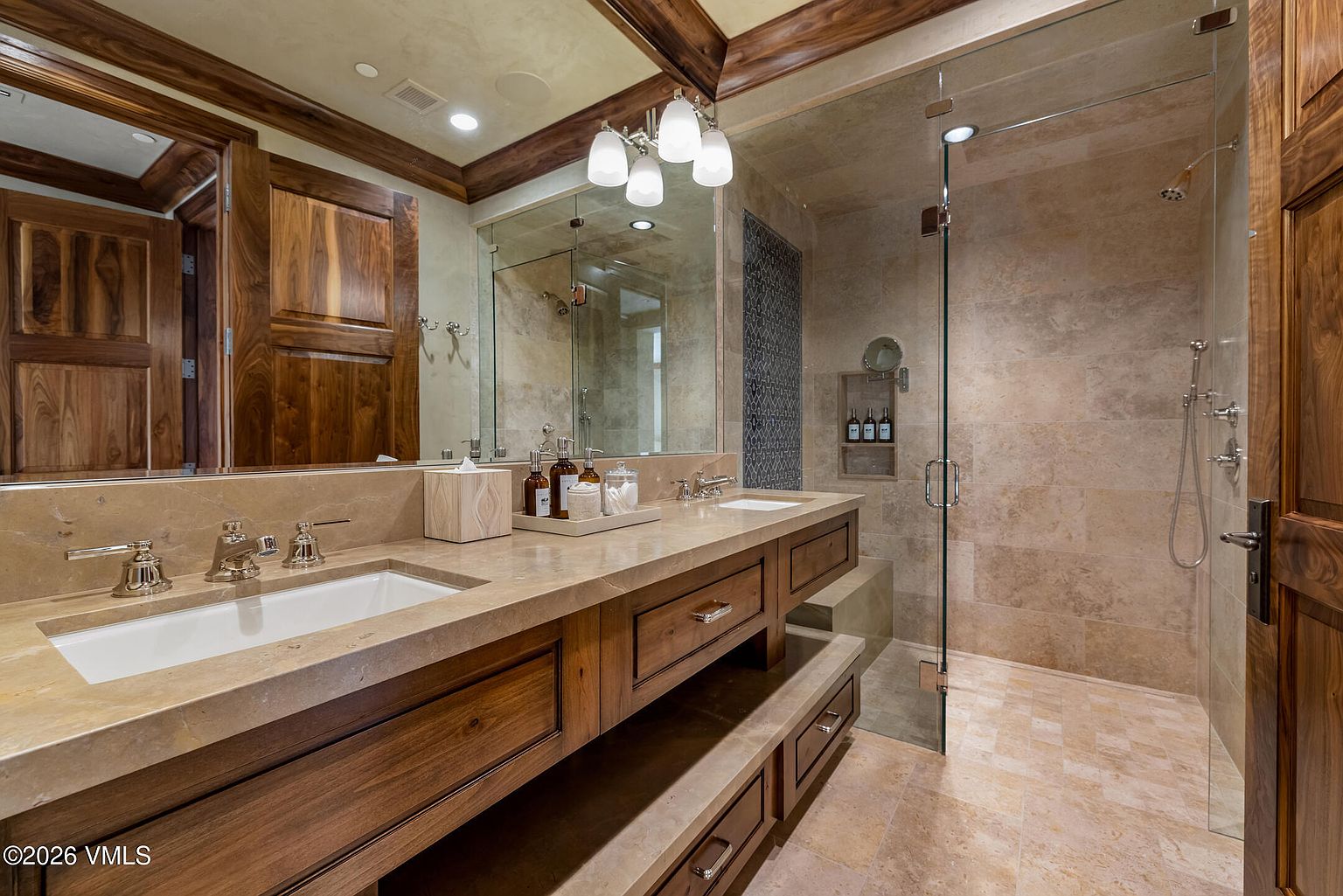 This luxurious primary bathroom features a dual-sink vanity with a stone countertop and rich wood cabinetry, complemented by a large mirror and elegant light fixtures. To the right, a spacious walk-in shower is enclosed by glass panels and finished with neutral-toned stone tile. The overall aesthetic is warm and sophisticated, blending natural textures with high-end finishes.