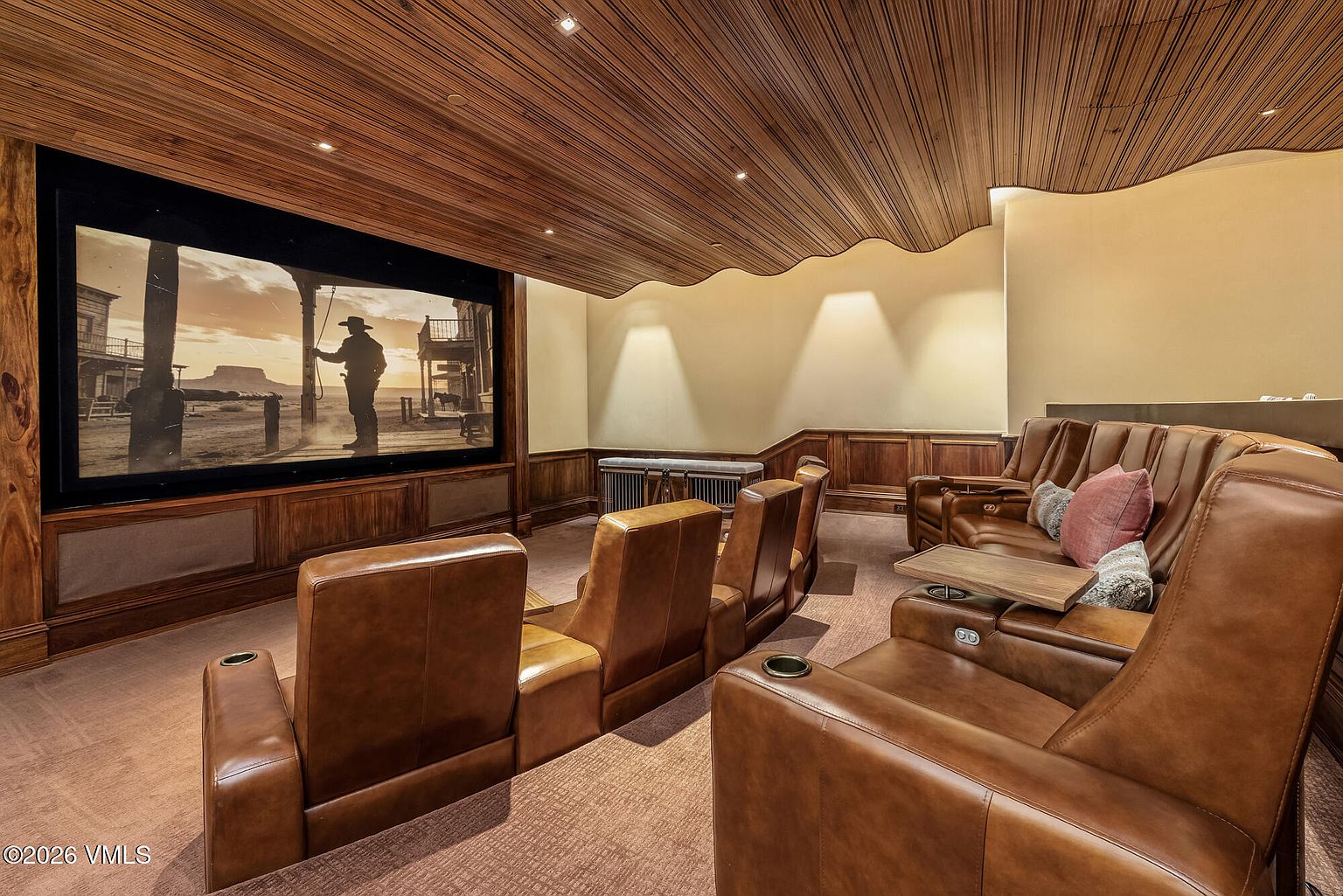 This dedicated home theater features tiered rows of plush, brown leather seating arranged for optimal viewing of a large, wall-mounted screen. The room is defined by its unique, undulating wood-paneled ceiling and warm, recessed lighting that creates an intimate, cinematic atmosphere. Wood wainscoting lines the lower walls, complementing the rich, warm tones of the carpet and furniture.