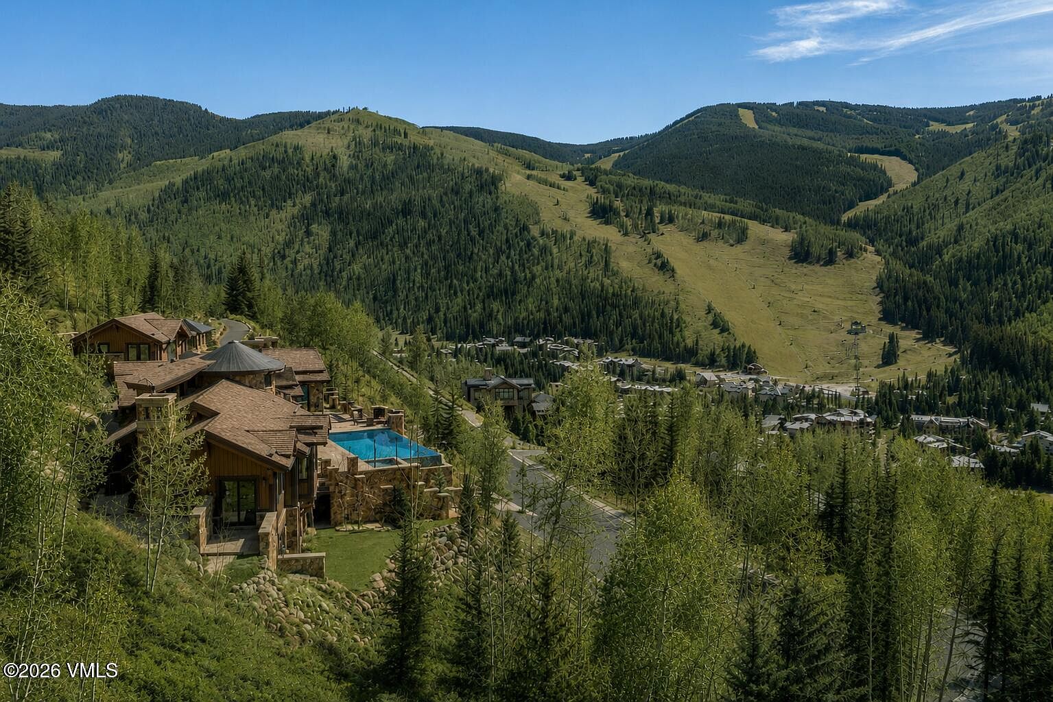 This high-angle aerial perspective captures a sprawling mountain estate nestled into a lush, forested hillside. The home features a multi-gabled roofline, stone exterior accents, and a prominent swimming pool overlooking a valley filled with dense trees and a distant residential community. The setting offers a serene, secluded atmosphere with expansive views of the surrounding ski slopes and natural landscape.