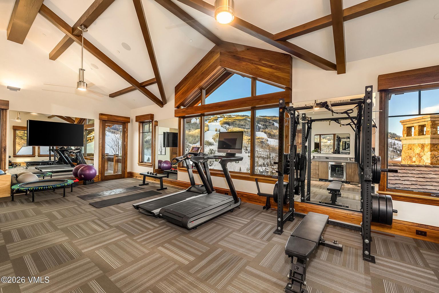 This spacious home gym features vaulted ceilings with exposed wooden beams and large windows that provide expansive views of a snowy mountain landscape. The room is equipped with a treadmill, a weight rack, and various fitness accessories, all set against a backdrop of mirrored walls and patterned carpet. A small kitchenette area with a beverage cooler is tucked into the corner, enhancing the convenience and luxury of this dedicated workout space.