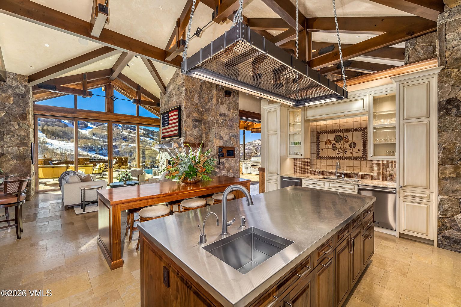 This expansive, rustic-style kitchen features a large central island with a stainless steel countertop and a professional-grade sink. The space is defined by heavy timber beams on the vaulted ceiling, a prominent stone-clad chimney breast, and a large wooden breakfast bar. Large windows provide a panoramic view of a snowy mountain landscape, creating a seamless connection between the luxurious interior and the outdoor environment.