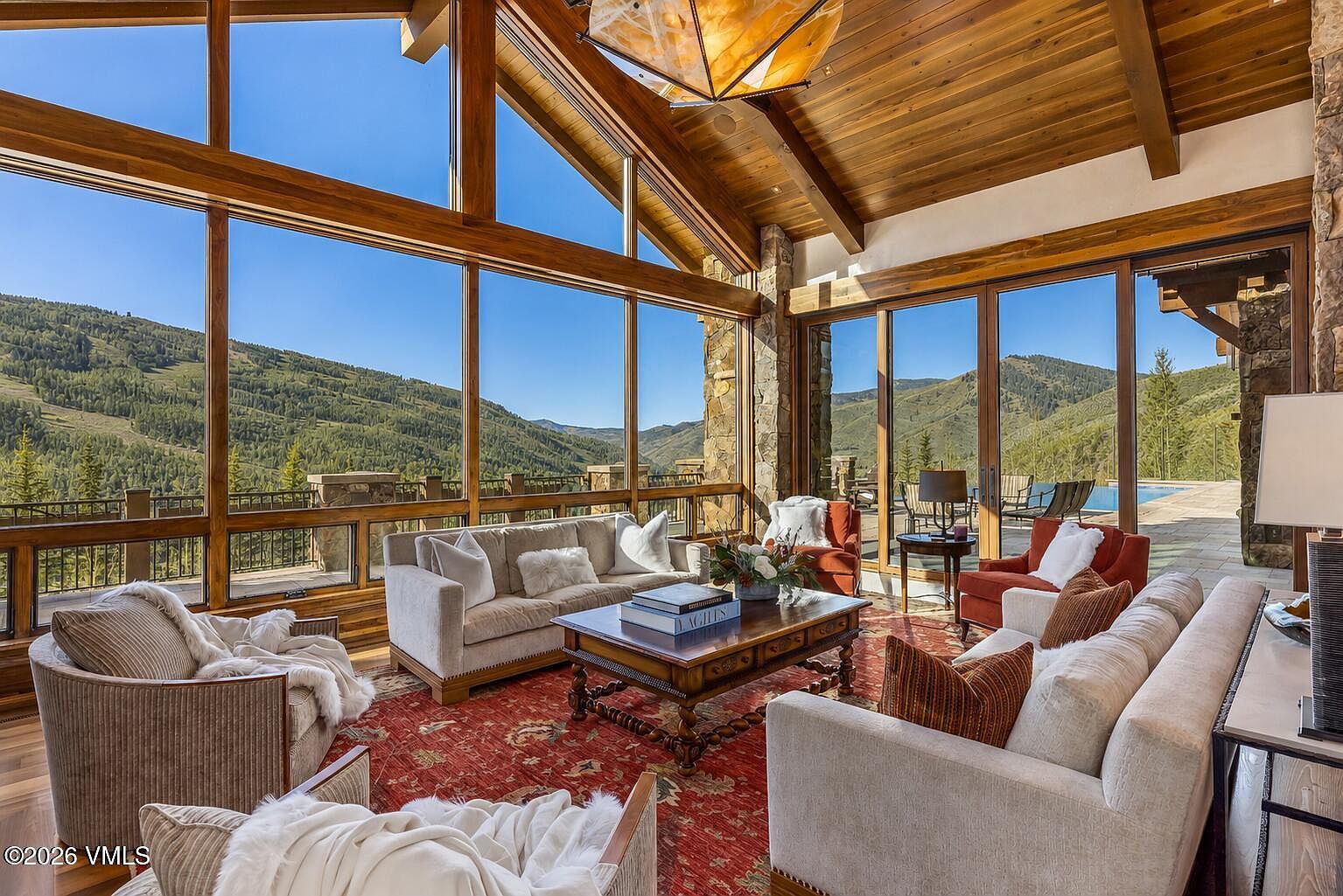 This expansive living room features a rustic, mountain-inspired aesthetic with a vaulted wood-paneled ceiling and large windows that frame panoramic mountain views. The space is furnished with plush, neutral-toned sofas and armchairs arranged around a central wooden coffee table, all set atop a vibrant red patterned area rug. The room seamlessly connects to an outdoor patio, creating an airy, open-concept feel that emphasizes the natural surroundings.