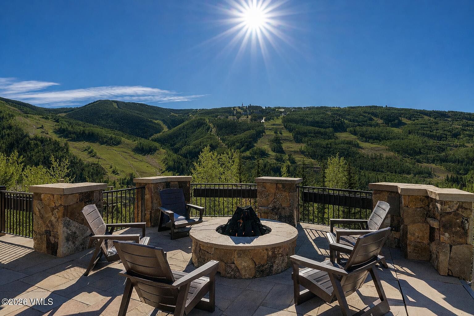 This expansive stone-paved patio offers a stunning elevated vantage point overlooking a lush, mountainous landscape under a bright, sunny sky. The space is centered around a circular stone fire pit surrounded by four dark wood Adirondack-style chairs, creating an inviting outdoor seating area. A sturdy stone and metal railing encloses the perimeter, emphasizing the dramatic, panoramic views of the forested slopes beyond.