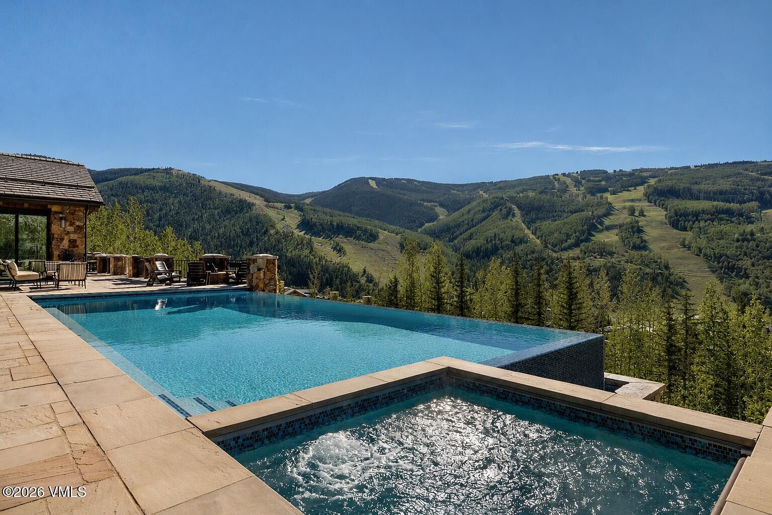 This stunning outdoor space features an infinity-edge pool and a separate hot tub, both set against a breathtaking panoramic view of a mountain ski resort. The stone-paved deck provides ample room for lounging, with outdoor furniture positioned to take full advantage of the expansive landscape. The scene captures a luxurious mountain retreat atmosphere, emphasizing the seamless connection between the home's amenities and the natural environment.