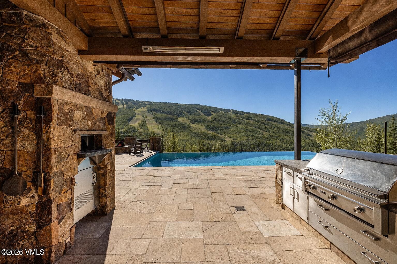 This expansive outdoor living space features a covered patio equipped with a built-in stainless steel grill and a stone-clad outdoor fireplace. The area transitions seamlessly to an infinity-edge pool that overlooks a scenic mountain landscape with ski slopes. The stone-paved deck provides a luxurious setting for entertaining, framed by a rustic timber-beam ceiling structure.