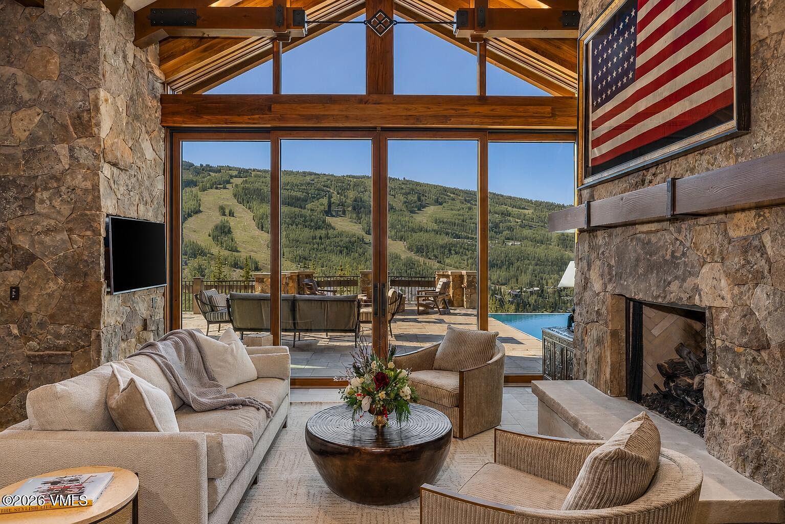 This expansive living room features a rustic, mountain-inspired aesthetic with a prominent stone-clad chimney breast and a large American flag displayed above the mantel. The space is anchored by a neutral-toned sofa and two armchairs arranged around a circular metal coffee table, all set against a backdrop of tall windows that frame a scenic mountain view. The room's vaulted timber-framed ceiling and stone walls create a warm, lodge-like atmosphere that seamlessly connects to an outdoor patio and pool area.