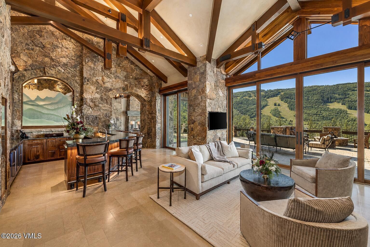 This expansive living area features a rustic mountain aesthetic with exposed timber beams and extensive stone wall accents. The space includes a built-in bar area with stools and a comfortable seating arrangement centered around a coffee table, all oriented toward large glass doors that reveal a scenic mountain view. The open-concept design seamlessly connects the indoor lounge to an outdoor patio, creating a bright and airy atmosphere.