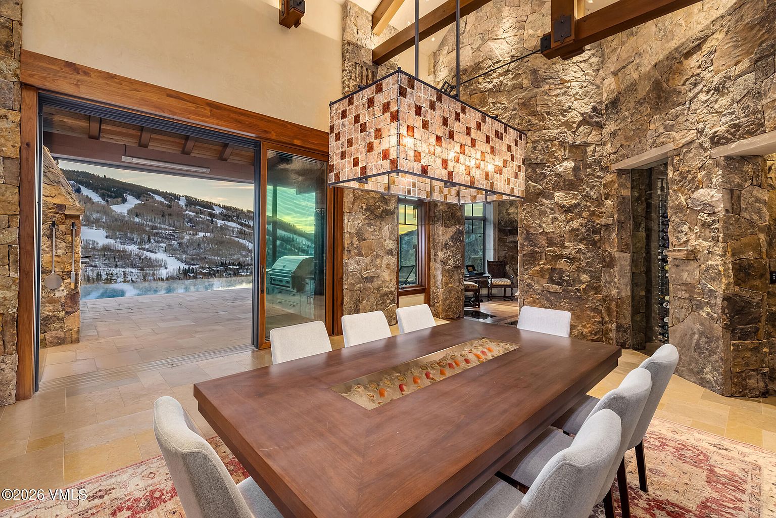 This sophisticated dining room features a large wooden table with a central integrated serving trough, surrounded by light-colored upholstered chairs. The space is defined by rugged stone walls and exposed timber ceiling beams, with a large, textured pendant light fixture hanging above the table. A wide opening leads to an outdoor patio with a scenic mountain view, creating a seamless transition between the indoor dining area and the exterior landscape.