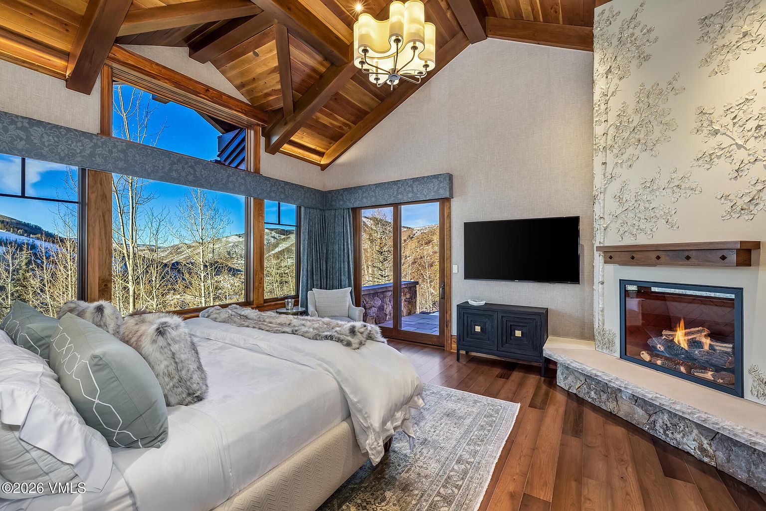 This luxurious primary bedroom features a vaulted wood-paneled ceiling with exposed beams and a large stone-clad chimney breast adorned with a decorative relief pattern above the mantel. A plush bed with textured pillows sits center stage, positioned to take advantage of the expansive mountain views through large windows. The room is completed by a dark wood media console, a stone hearth fireplace, and a neutral area rug, creating a warm and sophisticated mountain retreat atmosphere.