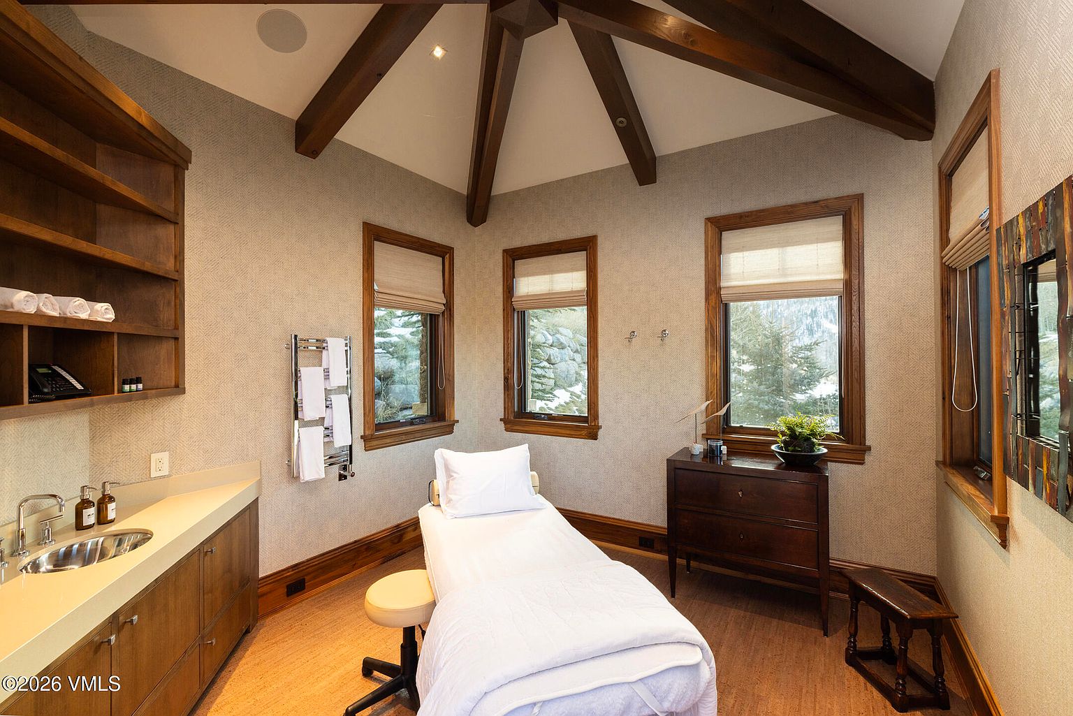 This professional spa or massage room features a central treatment table, a small stool, and a vanity area with a sink and storage cabinetry. The room is accented by exposed wooden ceiling beams, textured wall coverings, and three windows that provide natural light and views of the outdoors. The space is designed for relaxation, with a towel warmer and a dark wood dresser adding to the functional and serene atmosphere.