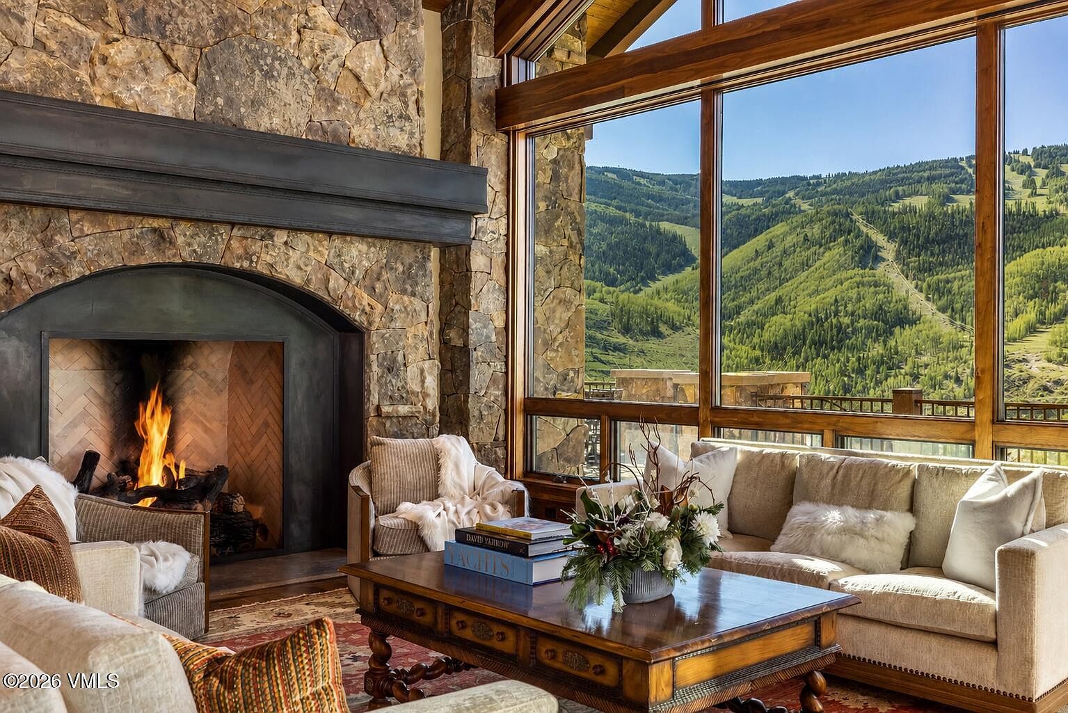 This inviting living room features a prominent stone-clad chimney breast with a large, arched fireplace as its focal point. Expansive windows frame a breathtaking view of the lush, mountainous landscape, bathing the room in natural light. The space is furnished with plush, neutral-toned seating and a substantial wooden coffee table, creating a warm and sophisticated mountain retreat atmosphere.