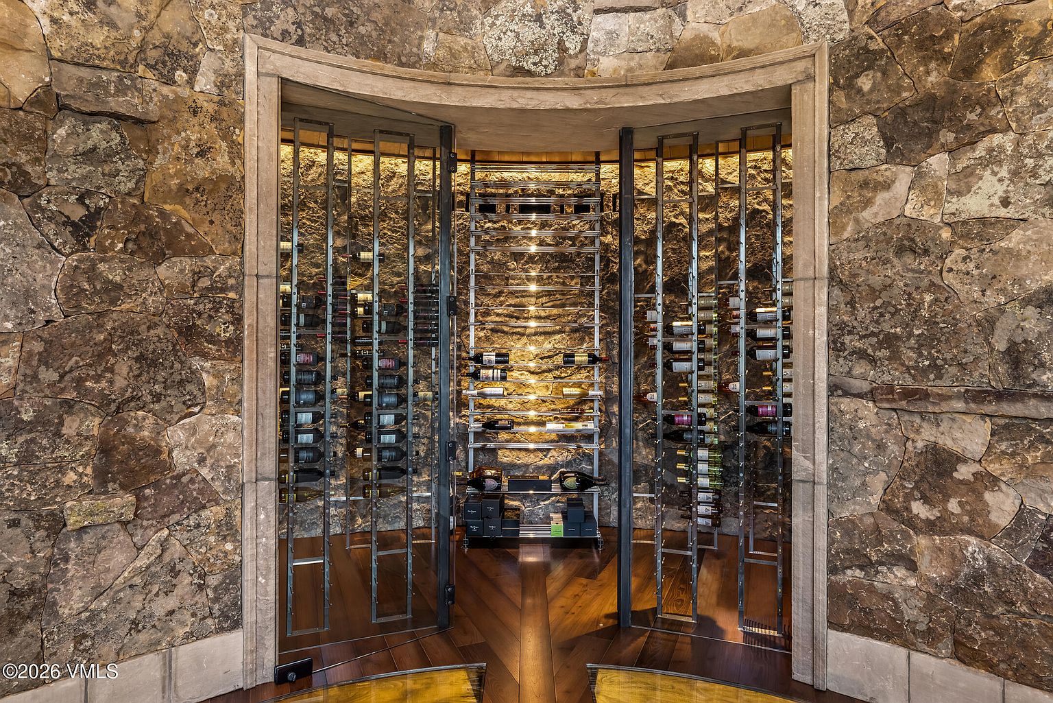 This sophisticated wine cellar features a curved, recessed design framed by a heavy stone archway. The interior walls are lined with rugged, natural stone, while a custom metal racking system provides organized storage for numerous wine bottles. The space is illuminated by warm, integrated lighting that highlights the texture of the stone and the collection, creating an inviting and luxurious atmosphere.