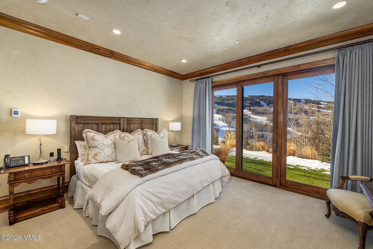 This primary bedroom features a warm, neutral color palette with textured walls and rich wood trim. A large bed with patterned pillows and a faux-fur throw serves as the focal point, flanked by two wooden nightstands with matching lamps. Large glass sliding doors provide an expansive view of a snowy, mountainous landscape, filling the room with natural light.