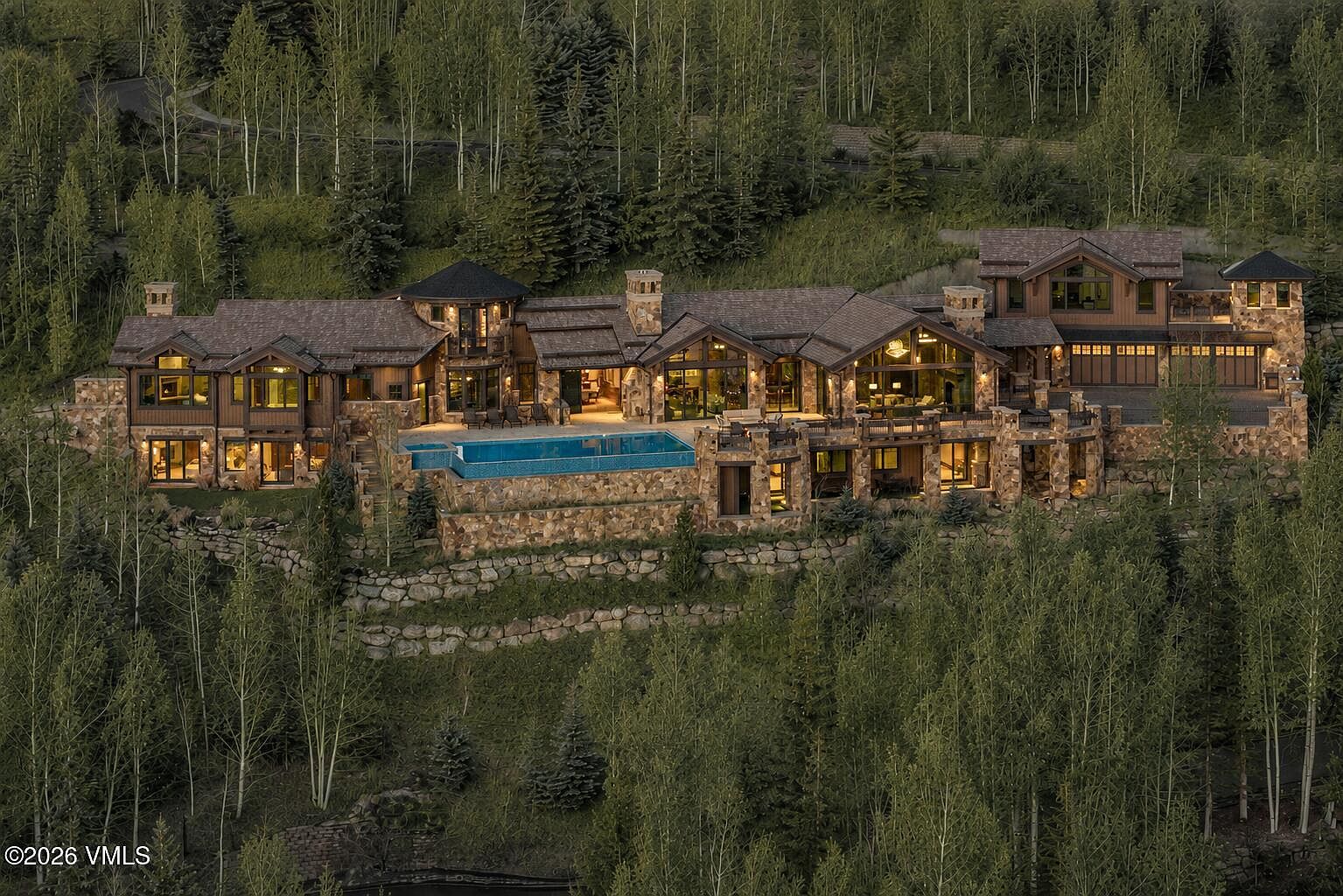 This expansive mountain estate is captured from an elevated aerial perspective, showcasing its sprawling, multi-level architecture nestled into a dense forest of aspen and pine trees. The home features a rustic design with stone masonry, dark wood siding, and multiple gabled rooflines, complemented by a prominent swimming pool overlooking the hillside. The composition emphasizes the property's secluded, high-altitude setting and its seamless integration with the surrounding natural landscape.