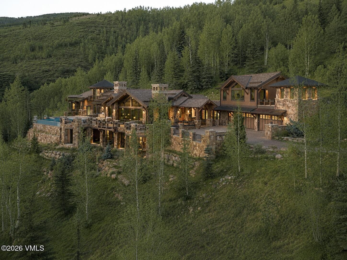 This expansive mountain estate is captured from an elevated perspective, showcasing a multi-level architectural design integrated into a lush, forested hillside. The home features a blend of stone and wood siding, with multiple gabled rooflines, a private swimming pool on the terrace, and a spacious paved motor court leading to a multi-car garage. The warm glow from the interior lighting contrasts beautifully against the twilight forest backdrop, emphasizing the property's secluded and luxurious character.