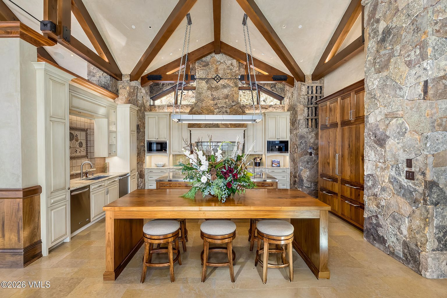 This expansive, rustic-inspired kitchen features a vaulted ceiling with exposed timber beams and a prominent stone-clad chimney breast that serves as a focal point. A large wooden island with three stools sits in the center, topped with a vibrant floral arrangement, while light-toned cabinetry and stainless steel appliances line the perimeter. The space is defined by its warm, natural textures, including stone walls and light-colored tile flooring, creating a grand yet inviting atmosphere.