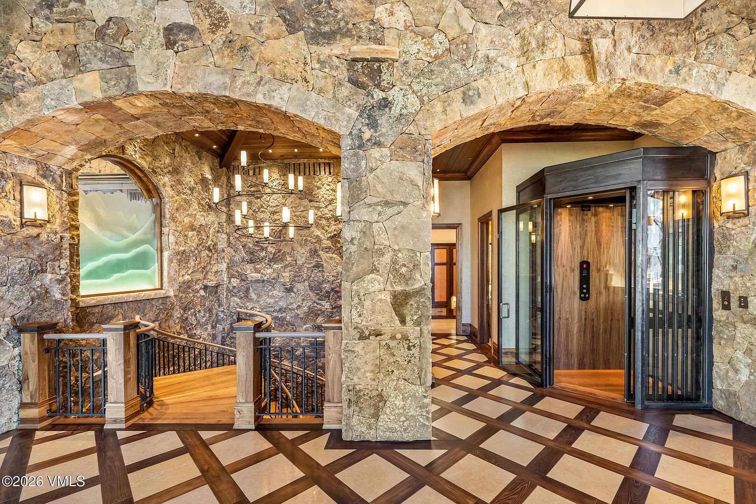 This grand foyer showcases a rustic, mountain-inspired aesthetic with heavy stone arches and a dramatic spiral staircase. The space features a sophisticated patterned tile floor, a custom glass-enclosed elevator, and a multi-tiered chandelier that adds a touch of elegance to the rugged stone walls. The perspective captures the transition between the stairwell and the main hallway, emphasizing the high-end craftsmanship and architectural depth of the home.